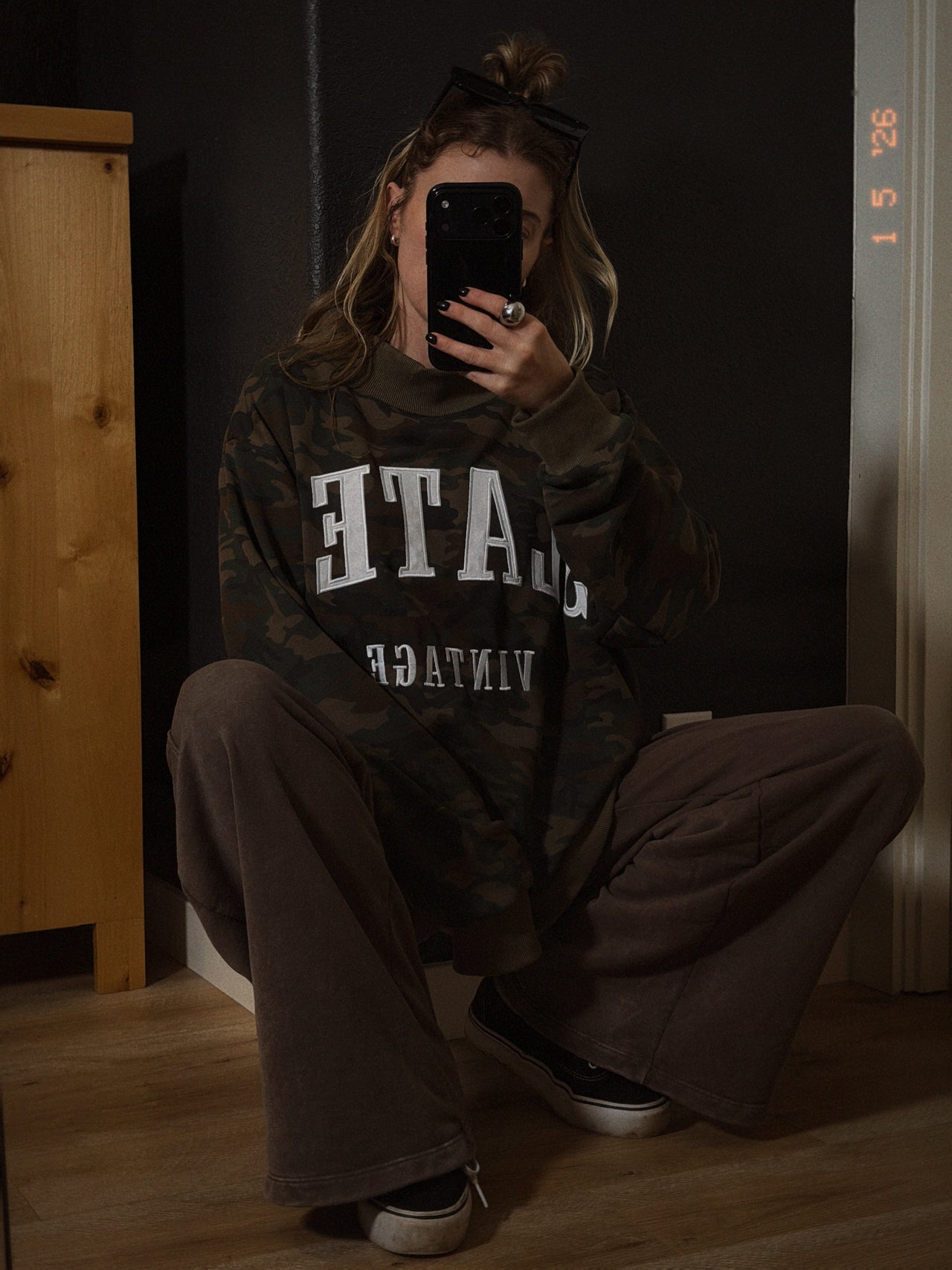 Person wearing a dark hoodie with visible text, taking a mirror selfie.