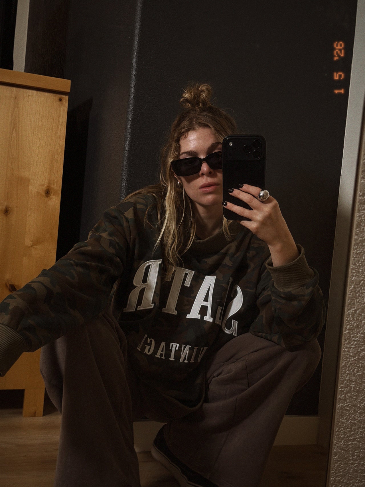 Person wearing a dark hoodie with visible text, taking a mirror selfie.