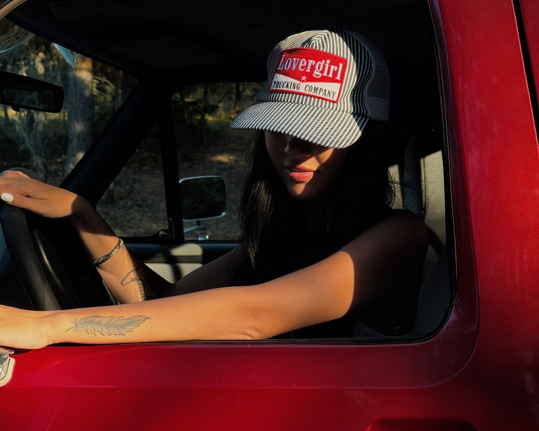 A woman sitting inside a red vehicle wearing a black and white striped trucker hat with a red and white 'lovergirl' patch.