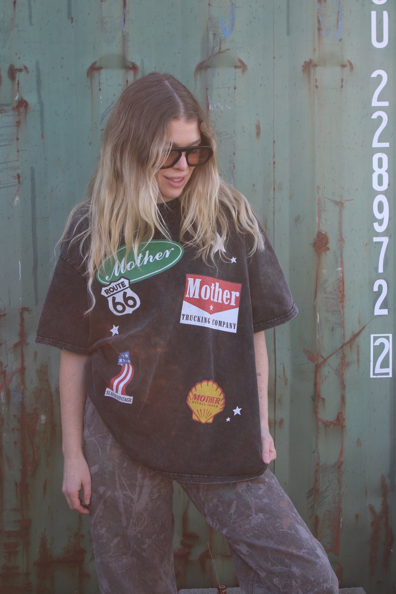 Person wearing a black t-shirt with various patches against a rusty metal wall.