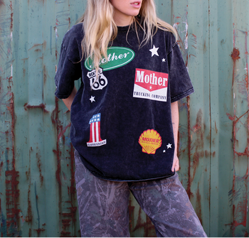 Person wearing a black t-shirt with various patches against a rusty metal wall.