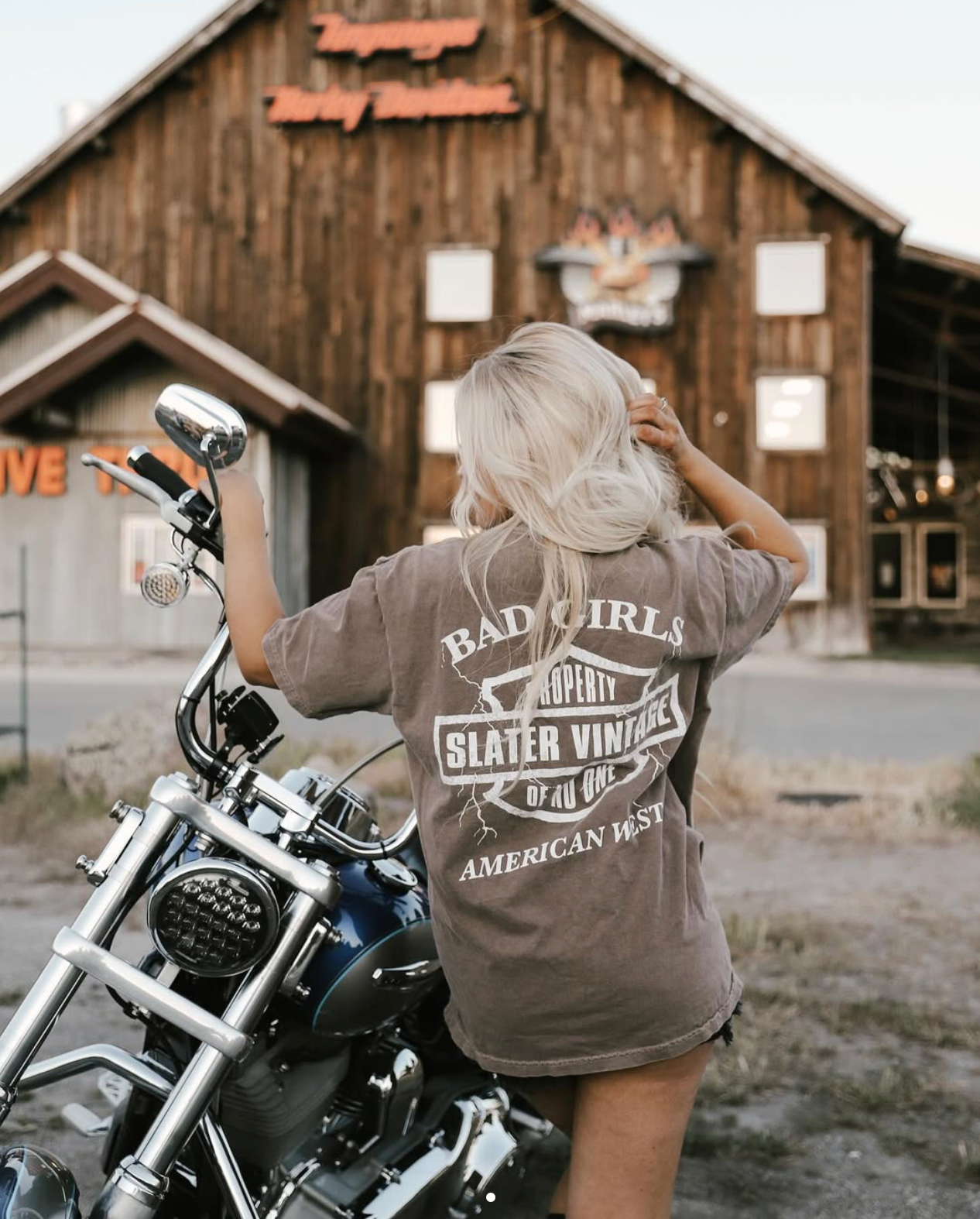 A blonde haired woman wearing a 'Bad Girls' t-shirt leaning against a motorcycle in front of a wooden building.